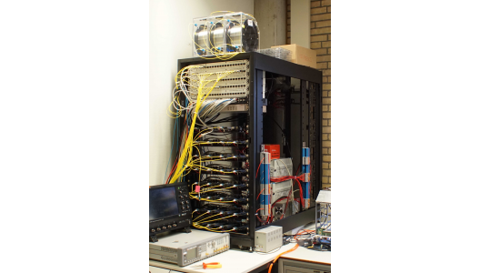 Figure 1. Timing System Setup in Amsterdam. It consists of several White Rabbit Switches from Seven Solutions (top panels in the rack), a copper ethernet switch, an Arbiter 1084B GPS, 32x ZEN-CTA boards (four in each of the lower panels of the rack) and several kilometers of single-mode optical fibers (e.g., on top of the rack).  Image credit: Heike Prokoph.